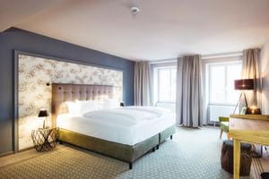 Moderne Hotel-Suite mit Doppelbett, grauem Polsterkopfteil, gemusterter Tapeten-Akzentwand, Holzschreibtisch und geometrischem Teppich.