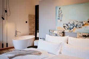 Modernes Hotelzimmer mit weißem Bett, freistehender Badewanne, künstlerischem Kopfteil und Pendelleuchten im Landhaus zu Appesbach.