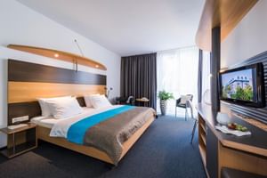 Helles Hotelzimmer mit Doppelbett, blauem Akzent, dunklem Teppich, moderner Beleuchtung, Sitzbereich und TV-Einheit. Große Fenster mit Vorhängen.