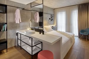 Modernes Smart-Doppelzimmer mit offenem Baddesign, weißem Bett, Betonwänden, schwarzen Metallarmaturen und beigen Vorhängen.