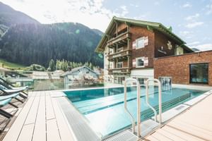 Piscine extérieure avec terrasse en bois à l'hôtel Schwarzer Adler à St. Anton, entourée de bâtiments alpins traditionnels et montagnes.