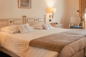 Elegantes Hotelzimmer mit Doppelbett, weißer Bettwäsche, dekorativem Kopfteil, Wandleuchte, Nachttisch, Polsterstuhl und goldenen Vorhängen.