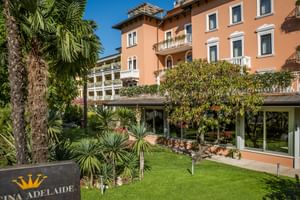 Elegantes korallfarbenes Hotel Regina Adelaide mit weißen Balkonen, umgeben von Palmen und gepflegten Gärten unter blauem Himmel.