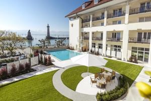 Garten des Hotel Reutemann mit Außenpool, kreisförmiger Rasenfläche mit Sitzbereich und Blick auf den Leuchtturm am Bodensee in Lindau.