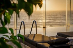 Sonnenuntergang-Terrasse im Macarico Beach Hotel mit Glasgeländer, dunklen Kissen und Meerblick. Grünes Laub rahmt den goldenen Horizont.