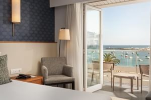 Modernes Hotelzimmer im Hotel Baia Cascais mit grauem Sessel, Balkon mit Blick auf Hafen mit Booten, Palmen und Meerblick in Portugal.