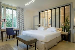 Helles Doppelzimmer mit weißem Bett, Glastrennwand zum Bad, Schreibtisch am Fenster mit gemusterten Vorhängen und moderner Beleuchtung im Auberge du Bon Laboureur.