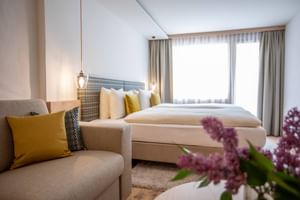 Bright hotel room with beige sofa, yellow cushions, double bed, patterned headboard, and purple flowers. Large window with curtains.