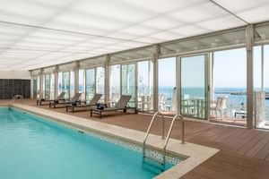 Innenpool im Hotel Baia Cascais mit bodentiefen Fenstern mit Meerblick. Poolbereich mit Liegestühlen und Holzboden.