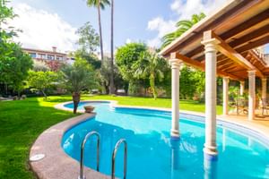 Piscine incurvée avec colonnes blanches sous terrasse couverte, entourée de pelouse verdoyante et palmiers à Son Sant Jordi à Pollença.