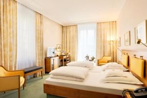 Modernes Doppelzimmer im Hotel Reutemann Lindau mit zwei Einzelbetten, Holzmöbeln, gelben Vorhängen, Schreibtisch und grünem Teppich.