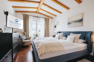 Modernes Doppelzimmer im Hotel Weisser Bär mit blau gepolstertem Bett, Holzdeckenbalken, dunkler Kommode und beigen Vorhängen.