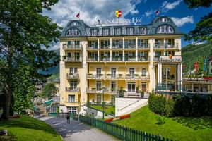 Gelbe Belle-Époque-Fassade des Hotel Salzburger Hof in Bad Gastein mit Balkonen, Mansardendach und Flaggen, umgeben von grünen Rasenflächen.