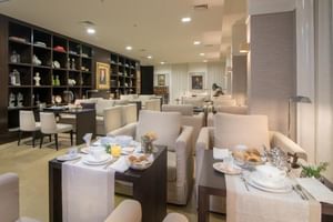 Moderner Frühstücksraum im Universal Boutique Hotel mit beigen Sesseln, gedeckten Tischen und dunklem Regal mit Dekorationsgegenständen.