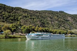 MS Switzerland Flusskreuzfahrtschiff fährt auf ruhigem grünen Wasser mit bewaldeten Hügeln im Hintergrund unter teilweise bewölktem Himmel.