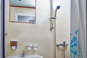 Kompaktes Badezimmer auf dem Schiff MS Macek mit weißem Waschbecken, Spiegel, Dusche mit Vorhang und Chromarmaturen an cremefarbenen Wänden.