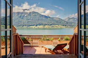 Holzbalkon mit Sonnenliege und Blick auf einen See und bewaldete Berge. Offene Balkontüren rahmen die malerische österreichische Alpenlandschaft.
