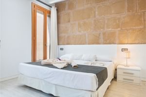 Modernes Doppelzimmer mit weißem Bett, Natursteinwand, Holztür, weißem Nachttisch mit Lampe und hellem Holzboden.