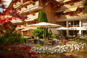 Wooden alpine hotel with flower-decorated balconies, garden terrace with white umbrellas, outdoor seating, and decorative pond in Gstaad.