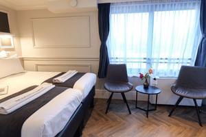 Moderne Juniorsuite auf MS MAGNIFIQUE IV mit zwei Einzelbetten, Sitzbereich mit zwei grauen Stühlen, kleinem Tisch mit Blumen und großem Fenster.