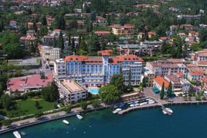 Luftaufnahme des Hotel Savoy Gardone, ein blau-oranges Hotel am Seeufer mit Pool, umgeben von mediterranen Gebäuden und Booten im Hafen.