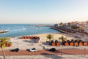 Sandstrand in Cascais mit orangen Strandhütten, Booten in der Bucht, Palmen und weißen Gebäuden entlang der Uferpromenade.