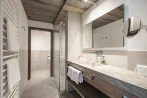 Salle de bain Rendl moderne avec poutres de plafond en bois, douche en verre, meuble-lavabo blanc avec plan en béton, grand miroir.