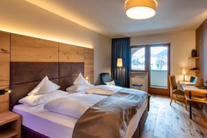 Modernes Hotelzimmer im Wiesnerhof Sterzing mit Holzkopfteil, weißer Bettwäsche, blauem Sessel, Schreibtisch und Bergblick-Fenster.