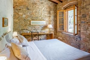 Rustikales Doppelzimmer mit Natursteinwänden, Holzbalken, weißer Bettwäsche, Schreibtisch mit Lampe und Rundbogenfenster mit Landblick.