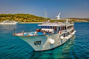 Weißes Kreuzfahrtschiff MY Mystic vor Anker in kristallklarem türkisem Wasser nahe einer bewaldeten Küste mit anderen Schiffen im Hintergrund.