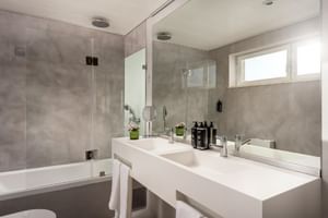 Modernes Badezimmer im Hotel Baia Cascais mit weißem Waschtisch, großem Spiegel, Badewanne mit Glasabtrennung und grauen Marmorwänden.