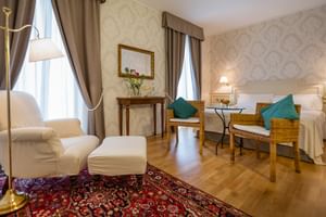 Elegantes Superior Doppelzimmer mit weißem Bett, cremefarbenem Sessel, Korbstühlen mit türkisen Kissen, Perserteppich und hohen Fenstern.