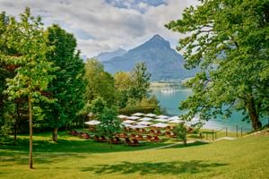 Seegarten im Landhaus zu Appesbach mit Liegestühlen und weißen Sonnenschirmen auf grüner Wiese, mit Blick auf türkisfarbenen See und Berge.