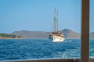 Weißes Segelschiff MS Macek mit zwei Masten auf blauem Wasser, mit felsigen Küsteninseln und Hügeln im Hintergrund unter klarem Himmel.