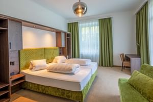 Modernes Doppelzimmer im Hotel Ertl mit grün gepolstertem Bett, eingebautem Kopfteil, Chrom-Pendelleuchte, grünen Vorhängen und passender Couch.