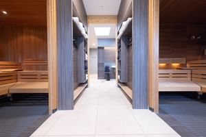 Heller Korridor zwischen zwei Saunaräumen mit Holzbänken und grauen Wänden. Weißer Fliesenboden führt zu einem Bereich mit Oberlicht.