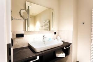 Modernes Hotelbad mit weißem Aufsatzwaschbecken auf dunklem Holzwaschtisch, großem Spiegel, Kosmetikspiegel und beigen Fliesen.