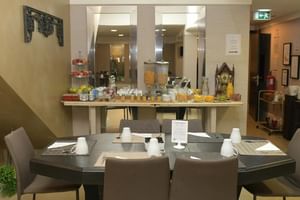 Moderner Frühstücksraum im Universal Boutique Hotel mit grauem Esstisch, beigen Stühlen und Buffetstation mit Getränken und Speisen.