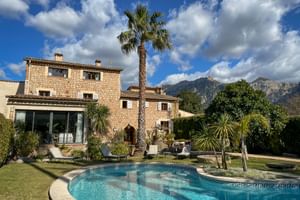 Hôtel Can Quatre en pierre à Sóller avec piscine extérieure, palmier et montagnes en arrière-plan sous ciel bleu et nuages blancs.