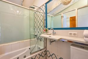 Salle de bain lumineuse avec baignoire blanche, paroi de douche en verre, miroir encadré bleu, meuble-lavabo blanc et suspensions tressées décoratives.