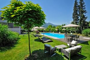 Garten vom Hotel Ertl in Spittal mit blauem Swimmingpool, Sonnenliegen, Gartenmöbeln und Sonnenschirmen auf grüner Wiese mit Bergen im Hintergrund.