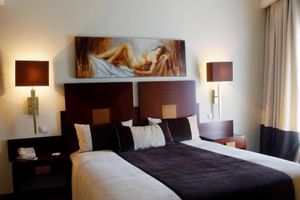 Modernes Doppelzimmer im Hotel Moliciero Aveiro mit dunklem Holzkopfteil, weißer Bettwäsche, Wandlampen und Kunstgemälde über dem Bett.