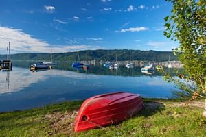 Rotes umgedrehtes Boot am Grasesufer des Bodensees mit Segelbooten im ruhigen blauen Wasser und bewaldeten Hügeln im Hintergrund.