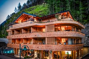 Bâtiment d'hôtel moderne incurvé avec façade en bois et éclairage chaleureux, niché contre une colline boisée dans les Alpes autrichiennes.