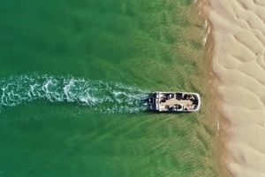 Luftaufnahme eines weißen Bootes mit Passagieren vor Faro Strand, das eine Bugwelle im grünen Wasser neben goldenem Sandstrand erzeugt.