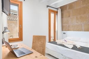 Modernes Hotelzimmer mit Natursteinwand, weißem Bett, Holzschreibtisch mit Laptop, Korbstuhl und Balkontür.