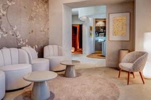 Elegante Spa-Lounge mit weißen geschwungenen Sesseln, runden Tischen auf beigem Teppich, floraler Wandkunst und Blick durch Bogen zum Saunaeingang.