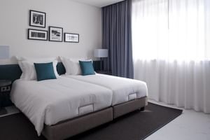 Modernes Hotel-Doppelzimmer mit zwei Einzelbetten, weißer Bettwäsche, türkisfarbenen Kissen und großem Fenster mit Vorhängen.