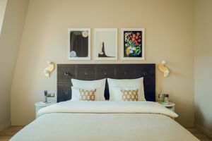 Modernes Hotelzimmer mit dunklem geometrischem Kopfteil, weißer Bettwäsche, Zierkissen, drei gerahmten Kunstwerken über dem Bett und Wandleuchten.