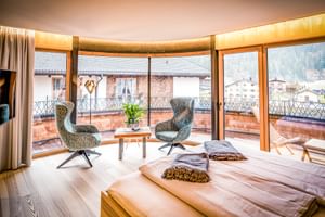 Chambre d'hôtel moderne au Hauserei Steeg avec baies vitrées courbes, deux fauteuils à motifs, balcon en bois et vue alpine.